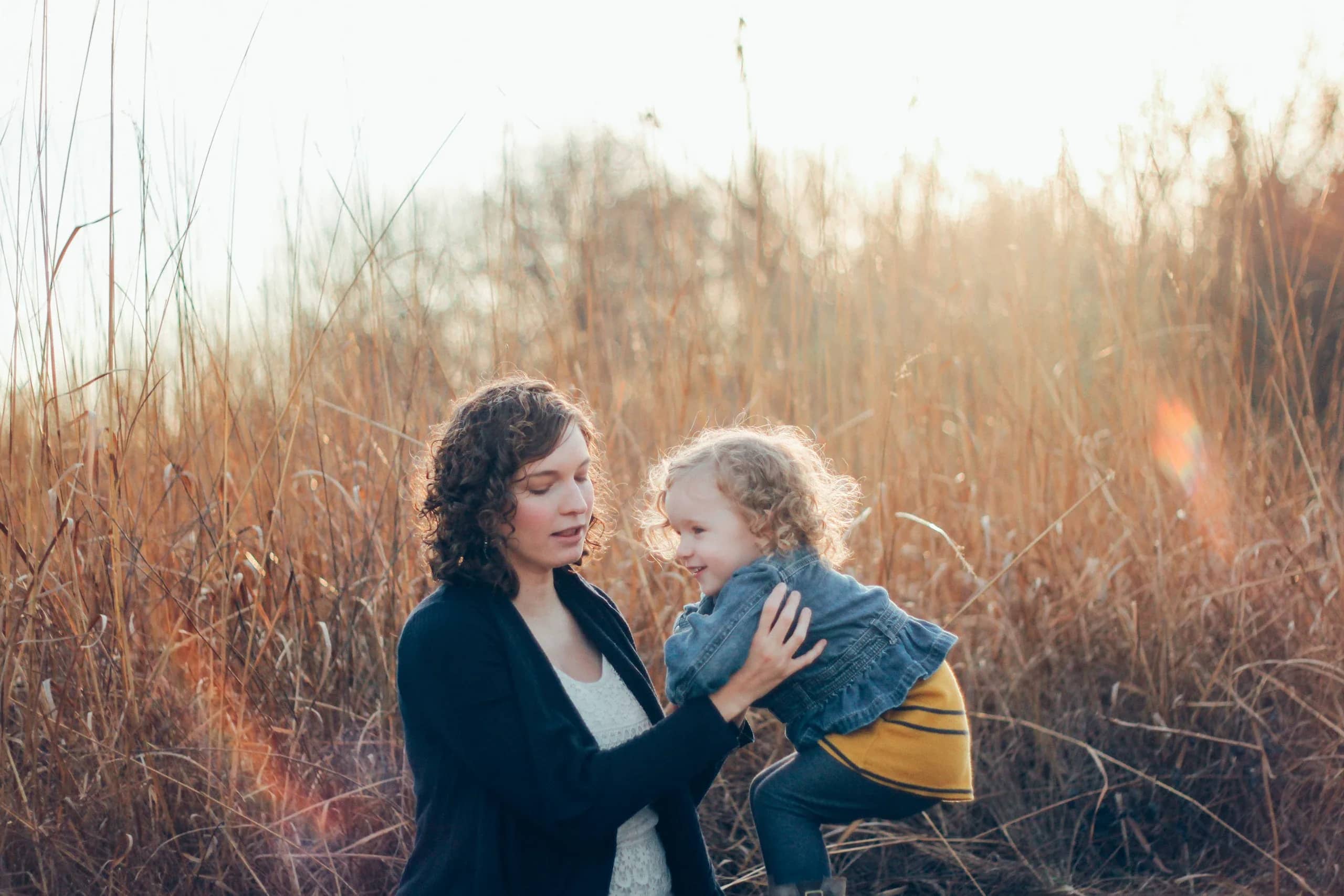 Mother lifting her laughing toddler in tall autumn grass, low sun behind them, cover of the Family & Newborn demo portfolio